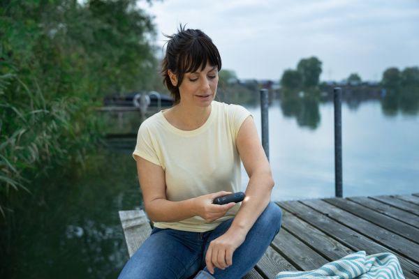 Eine Frau sitztin der Dämmerung auf einem Steg am Wasser und wendet den Insektenstichheiler BR 90 an.