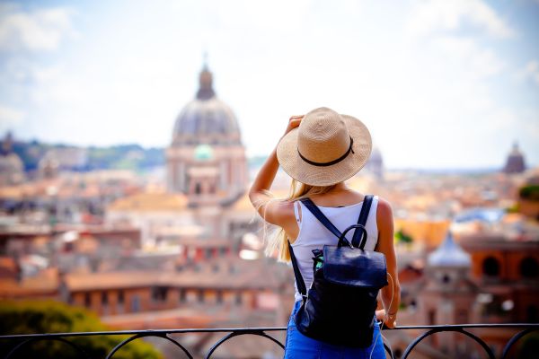 Eine Frau steht mit dem Rücken zur Kamera und trägt Sonnenhut und Rucksack. Sie blickt auf eine Dächerlandschaft, die mediterran wirkt.