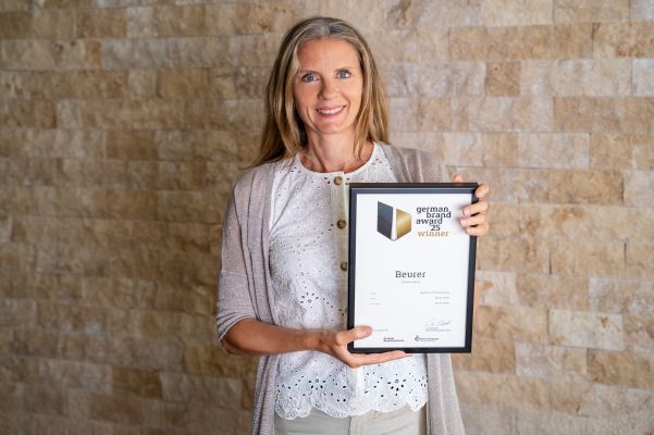 Beurer Marketingleiterin Kerstin Glanzer steht vor einer Steinwand und hält die German Brand Award Urkunde in der Hand.