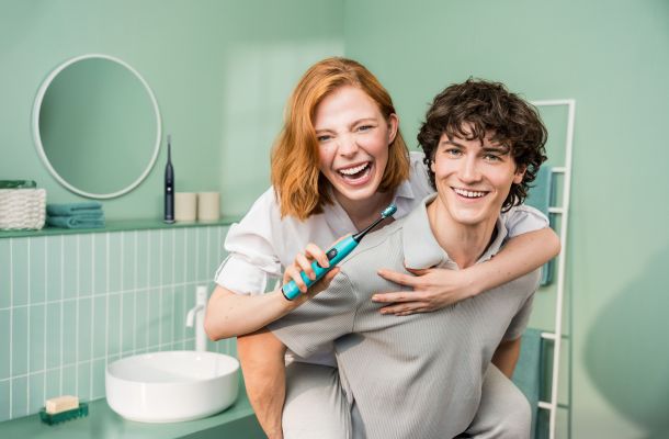 Ein junger Mann trägt eine junge Frau huckepack. Sie hält eine Beurer Schallzahnbürste und lächelt.