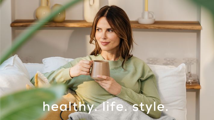 Eine dunkelhaarige Frau sitzt mit einer Heizdecke auf einem Sofa. Dazu der Beurer Markenclaim healthy. life. style. 