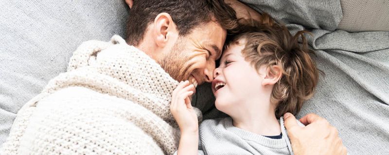 Vater und Sohn liegen Kopf an Kopf, eingekuschelt im Bett und lachen sich an
