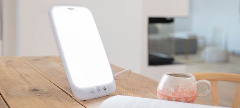 Die Beurer Tageslichtlampe TL 35 steht in einem Wohnzimmer auf einem Holztisch neben einer Tasse und einem Buch