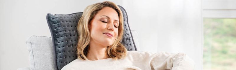 Frau sitzt im Sessel, lehnt sich an das Heizkissen HK 48 cosy und genießt mit geschlossenen Augen eine ruhige Pause