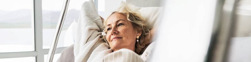 Ältere Frau sitzt in einem Sessel und blickt mit ruhigem Gesichtsausdruck aus dem Fenster