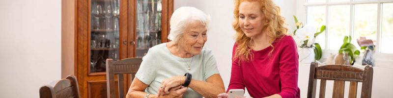 Ältere Frau mit Gardia Notrufarmband und jüngere Angehörige sitzen am Tisch, beide schauen auf ein Smartphone 