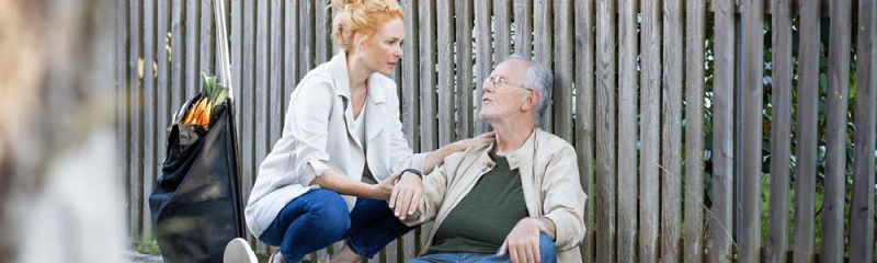 Älterer Mann mit Gardia Notrufarmband von Beurer sitzt draußen auf dem Boden am Zaun. Eine Frau kümmert sich um ihn