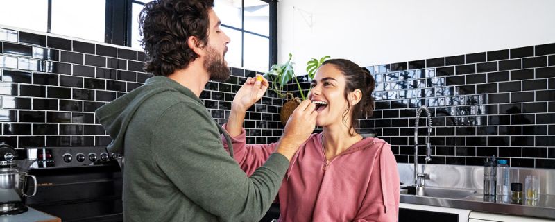 Ein Paar steht in der Küche, hält sich gegenseitig kleine Snacks hin und lacht dabei 