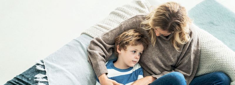Sohn liegt in den Armen seiner Mutter. Sie sitzen auf dem Sofa und die Mutter schaut zu dem Jungen hinunter.