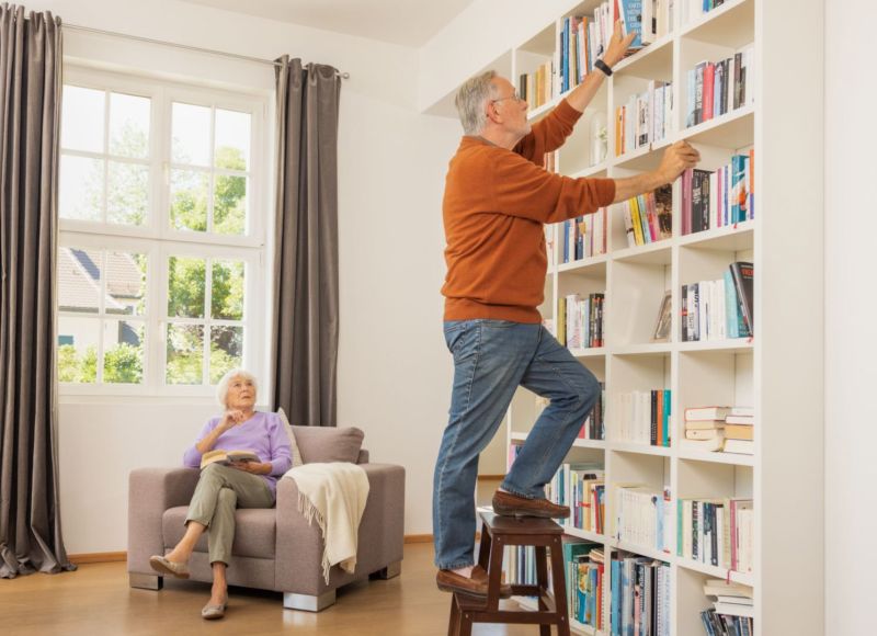 Älterer Mann mit Gardia Notrufarmband steht zuhause auf einer Trittleiter und greift nach Buch, Frau sitzt auf Sessel
