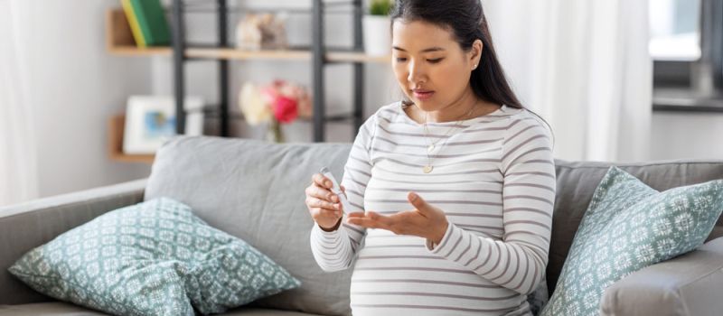 Schwangere Frau sitzt auf dem Sofa und misst ihren Blutzucker am Finger