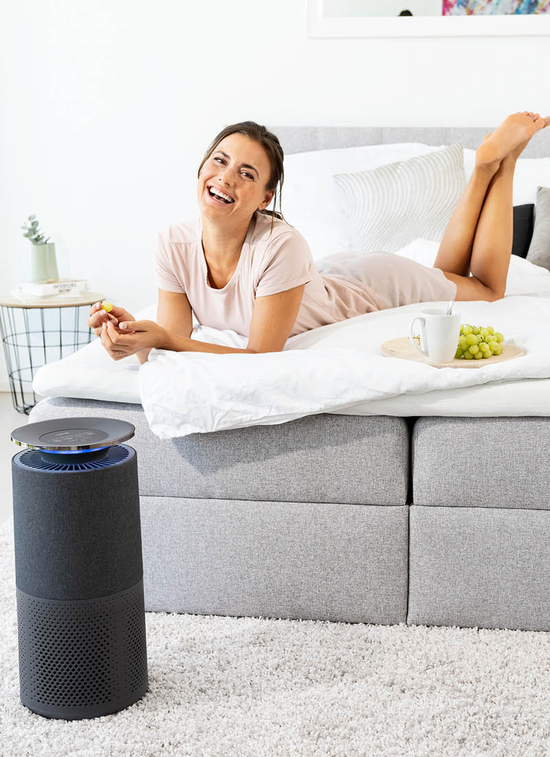 Junge Frau liegt lächelnd im Bett und Beurer Luftreiniger LR 401 steht im Vordergrund