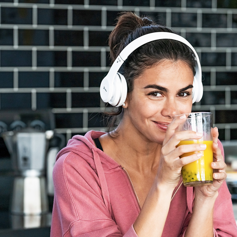 Eine Frau mit Kopfhörern genießt ein Glas Saft in der Küche und lächelt dabei entspannt in die Kamera