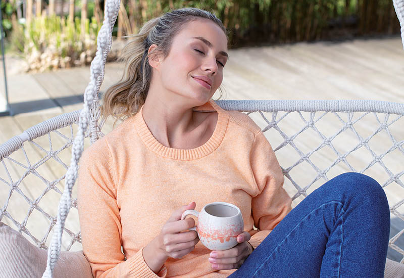 Junge Frau sitzt entspannt mit geschlossenen Augen in einem Hängesessel auf der Terrasse und hält eine Tasse 