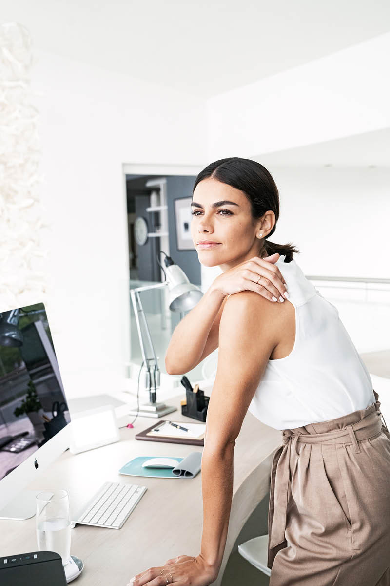 Eine junge Frau stützt sich auf ihrem Schreibtisch ab und hält sich an der Schulter