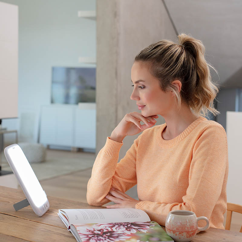 Frau sitzt am Tisch, blickt entspannt in die Beurer Tageslichtlampe TL 35, daneben liegt ein geöffnetes Magazin