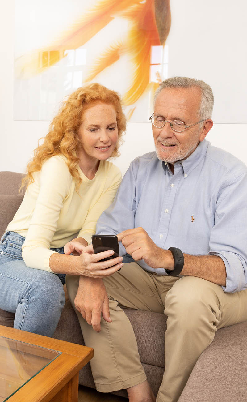 Älterer Mann mit Gardia Notrufarmband von Beurer lässt sich von Angehöriger die Funktionsweise am Smartphone zeigen