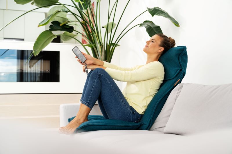 Junge Frau sitzt entspannt auf der Beurer Shiatsu-Sitzauflage MG 330 auf ihrem Sofa und hält den Bedienschalter in der Hand