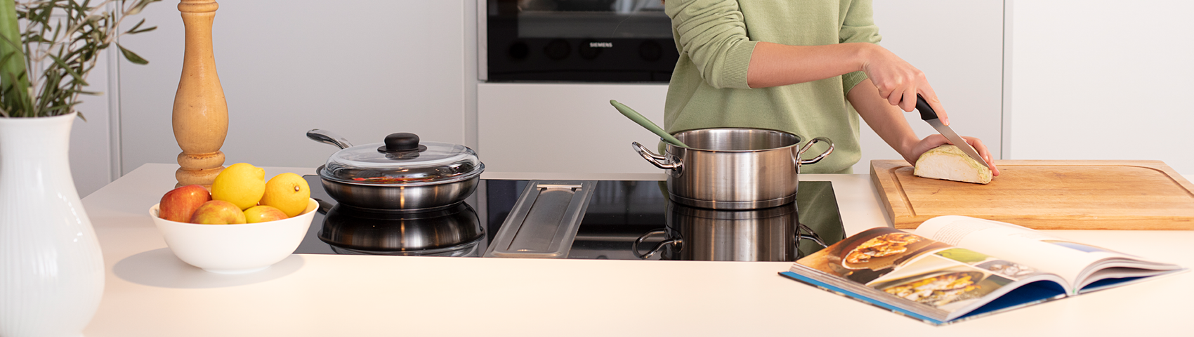 Lächelnde Frau hinter einem Herd schneidet Gemüse auf einem Brett und bereitet Essen mit Kochbuch zu
