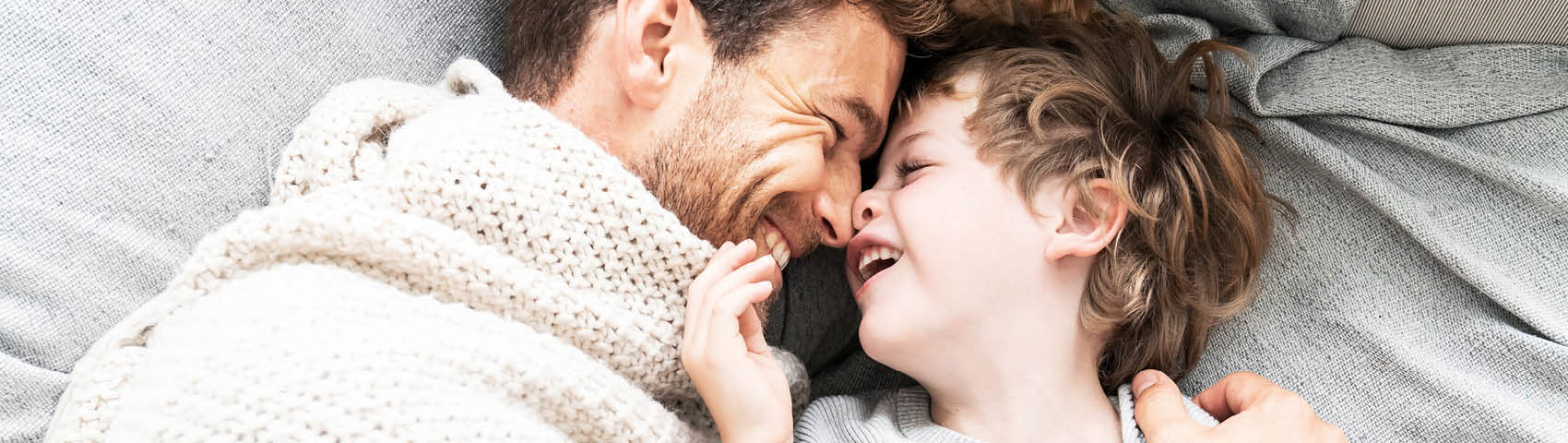 Vater und Sohn kuscheln unter einer Decke im Bett und schauen sich lachend an