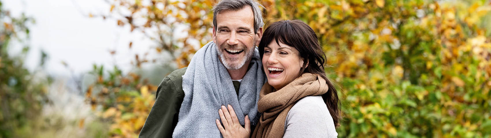 Ein warm angezogenes Paar mittleren Alters liegt sich lachend in den Armen draußen im Grünen