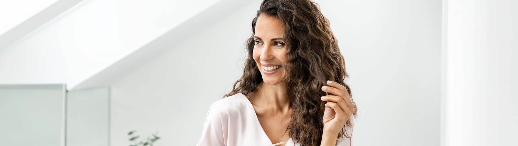 Frau mit langen, braunen, lockigen Haaren steht lächelnd im Bad und fährt sich durch die Haare