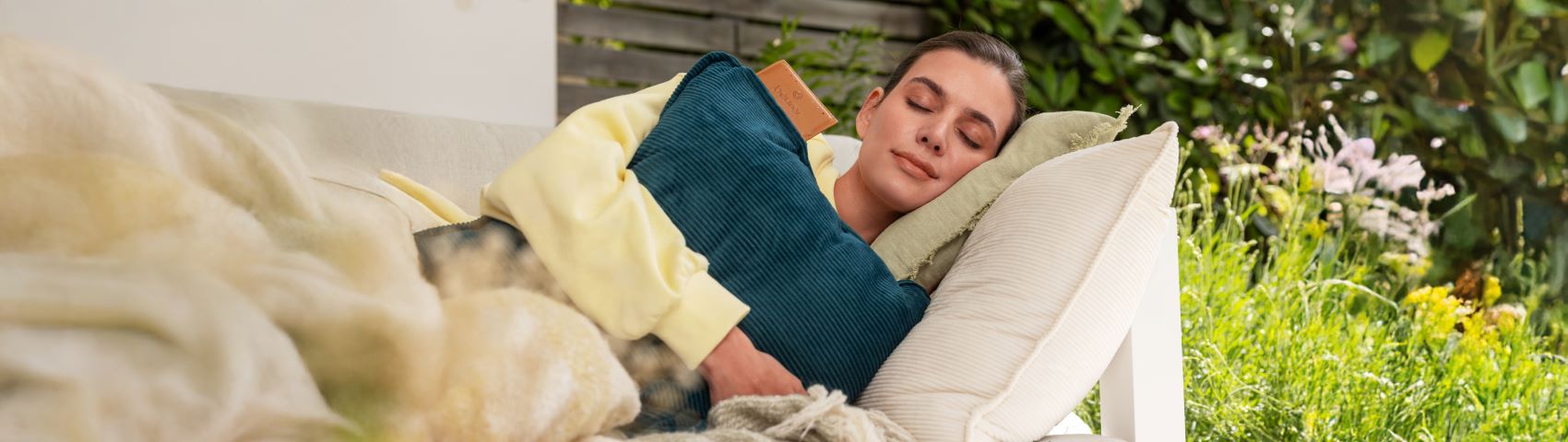 Junge Frau liegt mit geschlossenen Augen auf dem Sofa im Garten und hält das elektrische Heizkissen HK 77 von Beurer im Arm