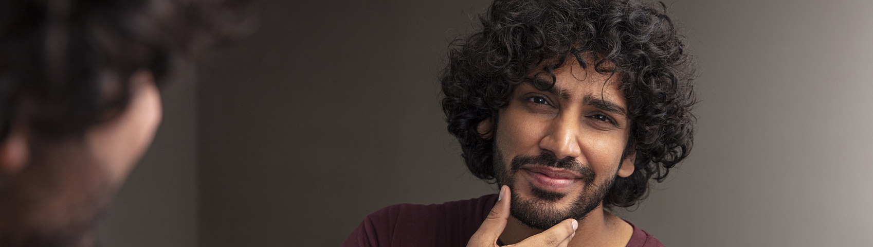 Mann mit dunklen, lockigen Haaren und vollem Bart schaut sich im Spiegel an und hält sich dabei das Kinn