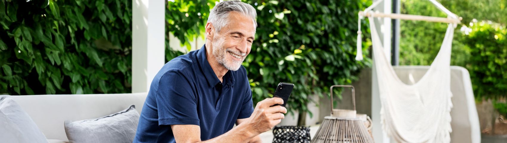 Älterer Mann mit grauen Haaren und Bart sitzt draußen auf einem Outdoor-Sofa und schaut auf sein Handy