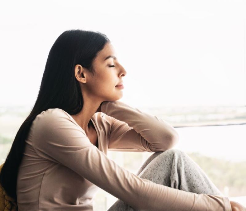 Junge Frau sitzt entspannt mit geschlossenen Augen und lächelnd an einem sonnigen Fenster