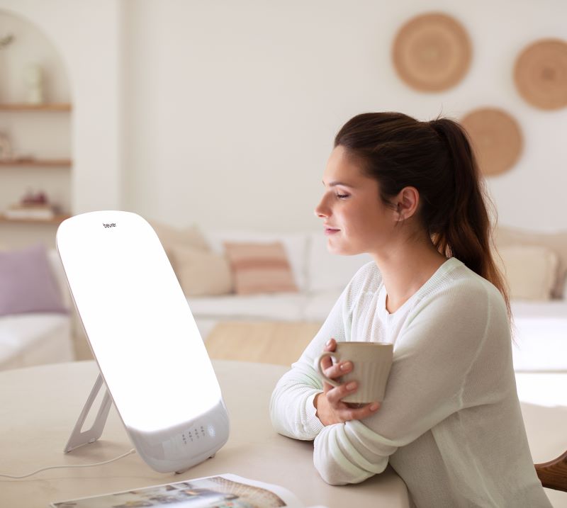 Junge Frau sitzt am Tisch mit einer Tasse in der Hand und nutzt die Beurer Tageslichtlampe TL 85