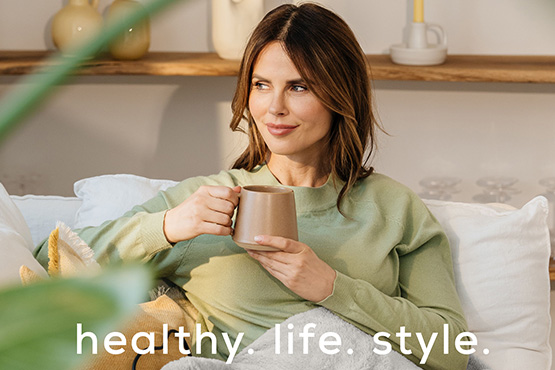 Eine dunkelhaarige Frau sitzt mit einer Heizdecke auf einem Sofa. Dazu der Beurer Markenclaim healthy. life. style. 