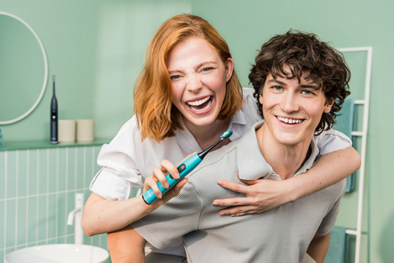 Ein junger Mann trägt eine junge Frau huckepack. Sie hält eine Beurer Schallzahnbürste und lächelt.